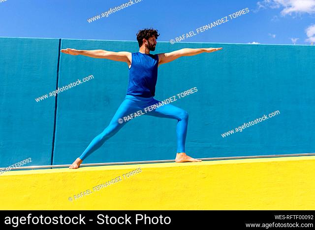 Mid adult man practicing Virabhadrasana II on retaining wall during sunny day