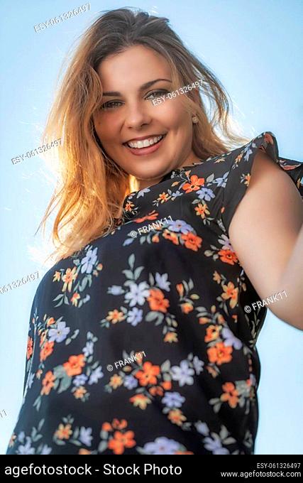Bottom view of attractive beautiful young woman standing outdoors against blue sky and smiling at the camera