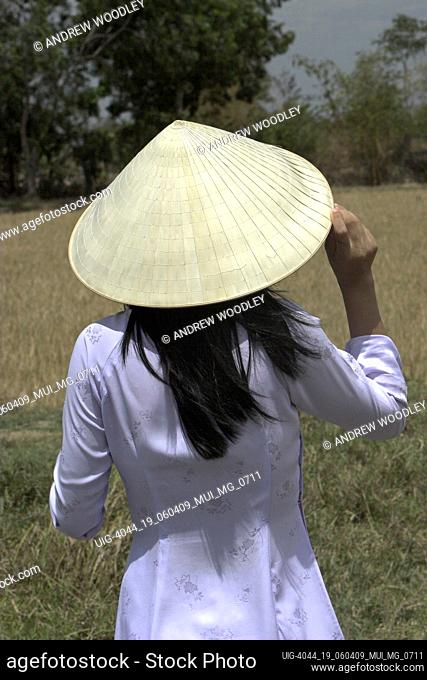 Young woman in traditional white ao dai holding conical hat on head looks across fields Vietnam