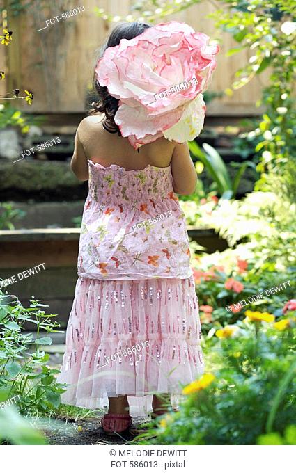 A Rear View of a young girl dressed up standing in a garden and carrying a paper flower