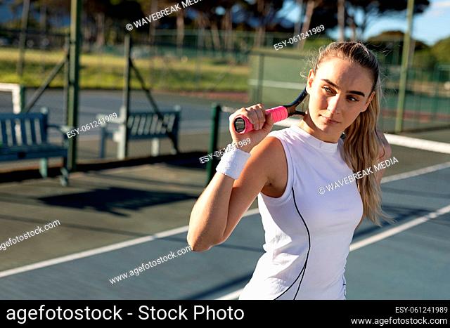 Confident young female caucasian tennis player with racket around neck at court on sunny day