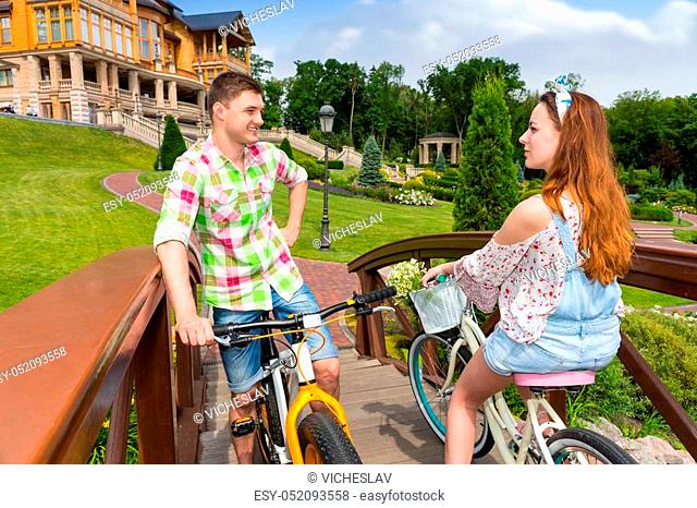 Meeting of a girl and guy in green and red plaid shirt while they are riding on bikes on footbridge in the beautiful park