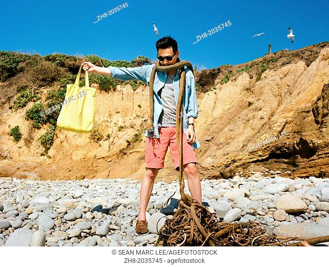 A man in summer beach wear wrapped with sea plants like a scarf while holding a bag out along the coast of Northern California's Pacific Coast Highway
