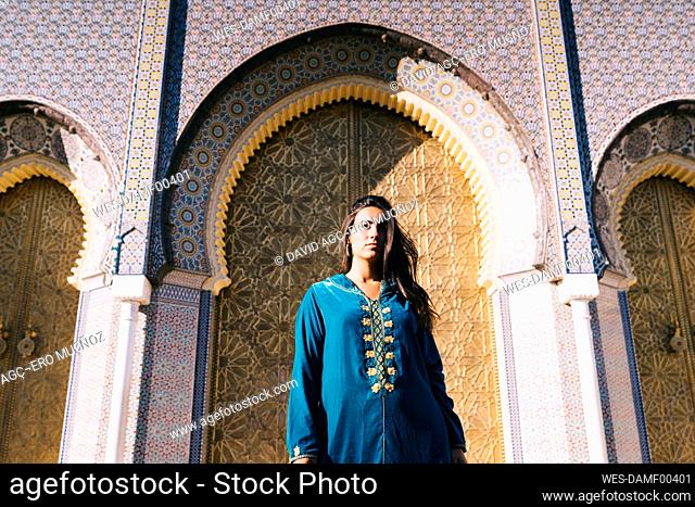 Young woman standing in front of traditional architure wearing Moroccan robe, Morocco