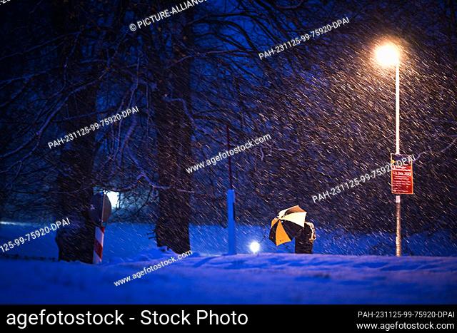 25 November 2023, Baden-Württemberg, Isny im Allgäu: Snowflakes are blowing around a street lamp at the blue hour as a woman with an umbrella walks past