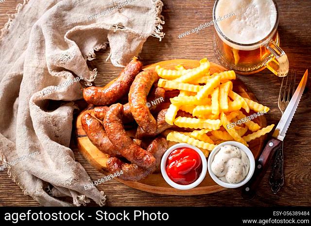 fried sausages with french fries on wooden board and mug of beer, top view