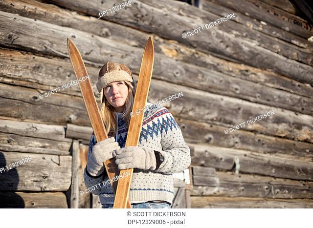 Portrait Of A Young Woman Holding Wooden Skis By A Log Cabin; Homer, Alaska, United States Of America