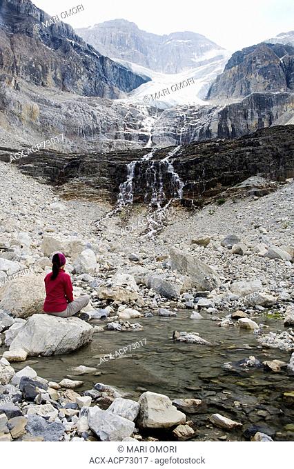 A hiker sits on a rock beside a pool of water fed by the Stanley Glacier. Smoke from nearby wildfire hang over the mountains. Model Release signed