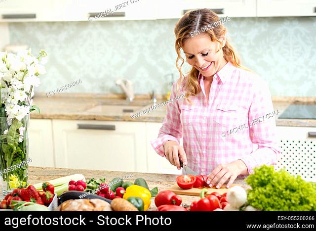 smiling woman in a shirt is slicing tomatoes on a kitchen table with vegetables