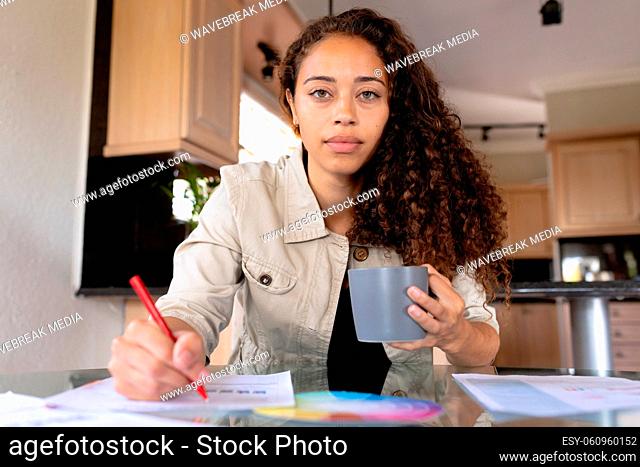 Portrait of confident biracial female freelancer holding cup working at home office