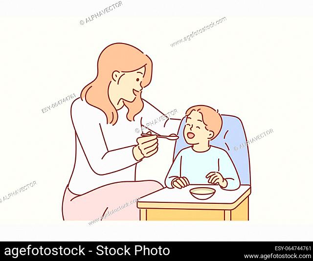 Mother feeds boy sitting in child seat with spoon, caring for baby and wanting to give son best. Woman works nanny in kindergarten and feeds baby with healthy...