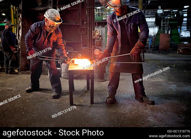 Forge worker hands over pre formed steel billet to colleague for presentation to set of two dies in hammer to be fully forged into crank shaft