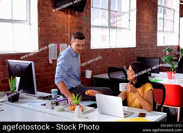 Caucasian male and female colleague sitting in office having coffee and pastries and laughing