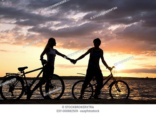 Silhouettes of couple holding each other by hands while walking down seashore with their bicycles at sunset