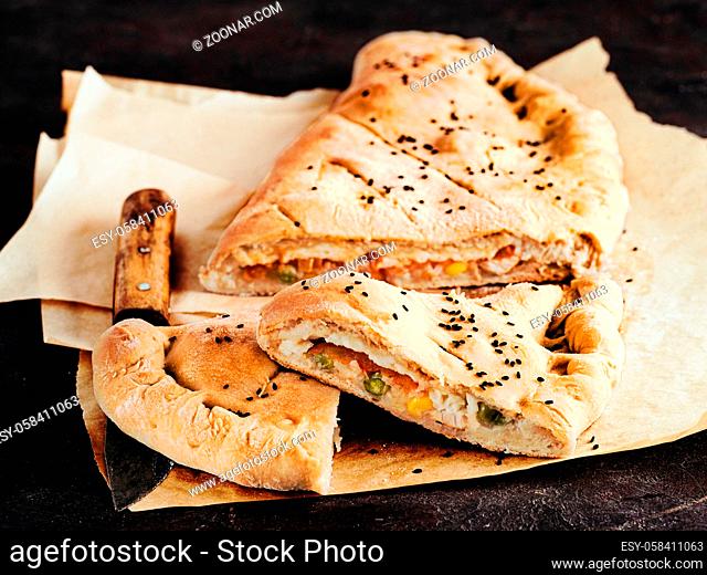 Perfect homemade closed pizza calzone on baking paper sheets over black background. Cutting tasty italian closed calzone pizza with cheese, meat, vegetables
