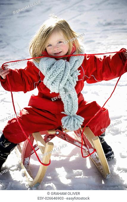 Little girl sitting on a wooden sled in the snow. She is smiling at the camera and holding the rope up from the sled