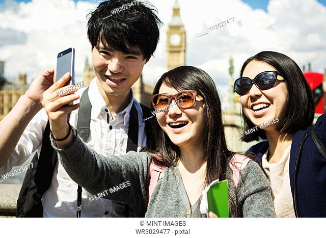 Smiling man and two women with black hair taking selfie with smartphone, standing on Westminster Bridge over the River Thames, London
