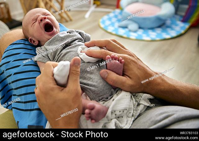 Father's hands holding three month old baby boy yawning on lap, indoors