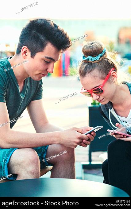 Couple of friends, teenage girl and boy, having fun using smartphones sitting in center of town, spending time together