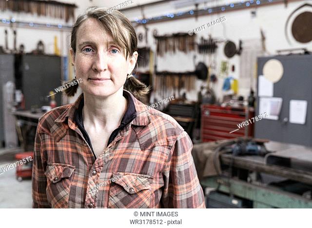 Blond woman wearing checked shirt standing in metal workshop, looking at camera