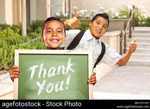 Happy hispanic boys giving thumbs up holding thank you chalk board outside on school campus