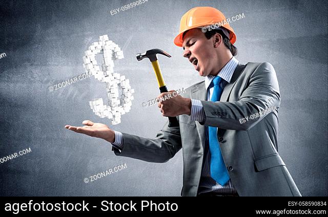 Furious businessman going to crash with hammer dollar symbol. Young handsome man in business suit and safety helmet standing on wall background