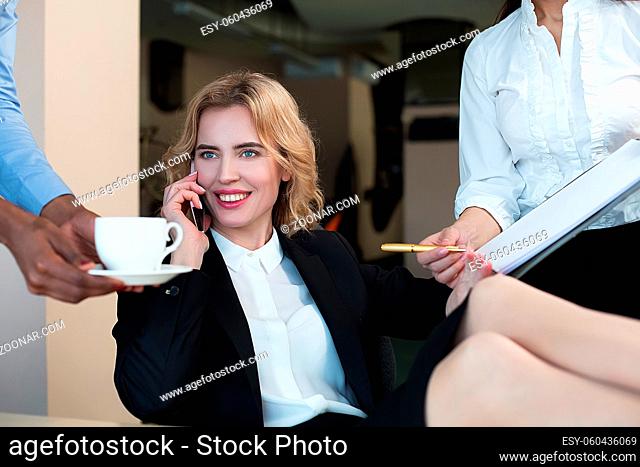 Smiling woman speaking over phone. Assistant brought coffee for her