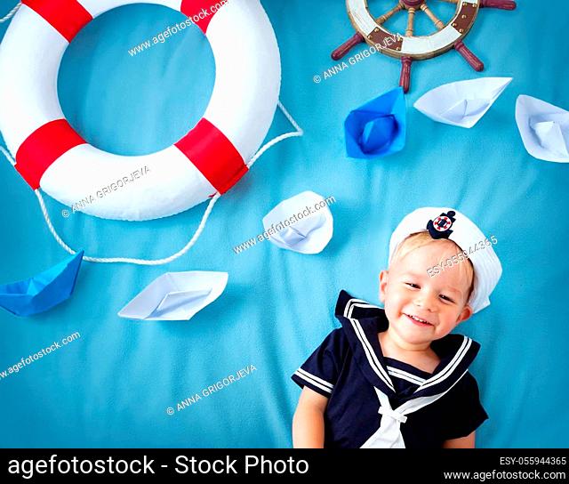 two years old boy playing in sailor hat. Child with a steering wheel and paper ships on blue background