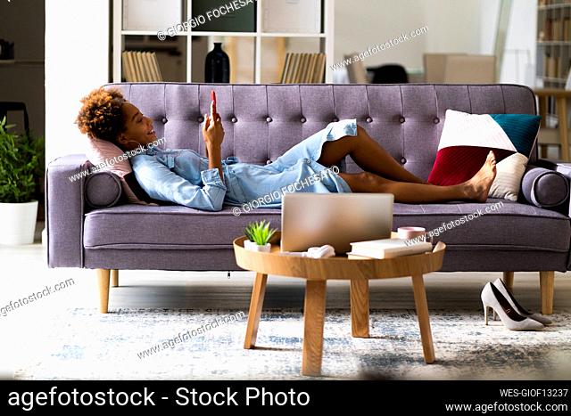 Smiling young woman using smart phone while lying on sofa at home