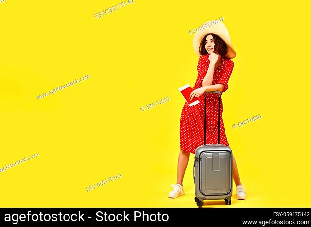 beautiful woman in red dress with gray suitcase and and passport going traveling on yellow background. copy space