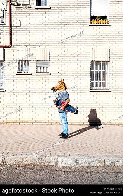 Man wearing horse mask holding skateboard on footpath by building in city during sunny day