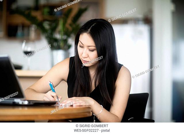 Young woman in black dress using laptop and taking notes on table at home