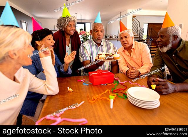 Multiracial senior friends wearing party hats looking at man blowing candles in nursing home