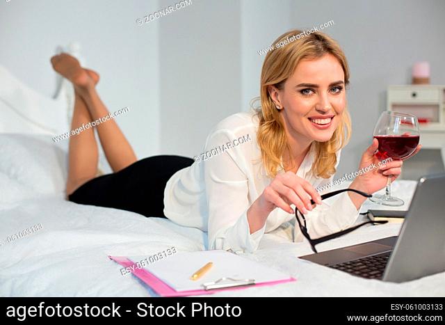 Smiling woman making business in bed and enjoying glass of wine. Isolated on white background