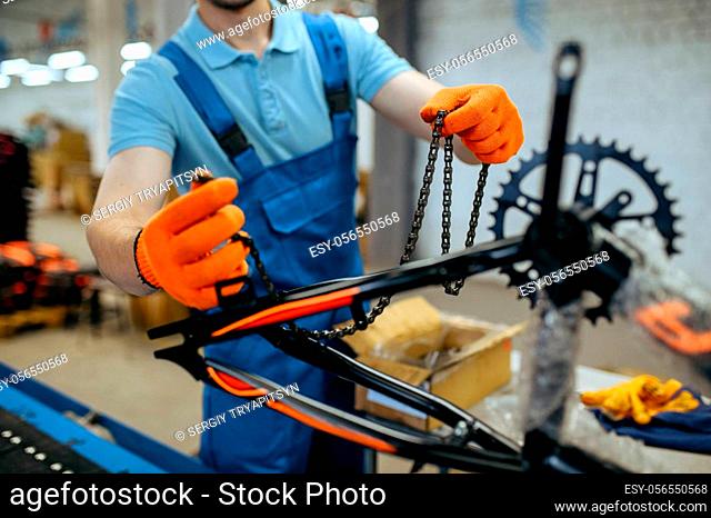 Bicycle factory, worker holds bike chain at assembly line. Male mechanic in uniform installs cycle parts in workshop, industrial manufacturing