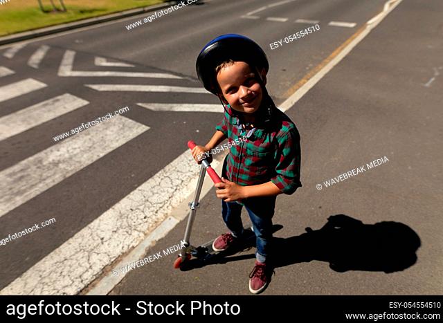 Portrait of a Caucasian schoolboy wearing a helmet and standing with a scooter smiling to camera while waiting at a pedestrian crossing to cross the road safely...