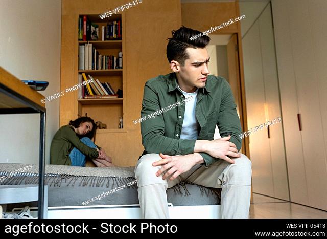 Sad young woman looking at displeased boyfriend sitting on bed at home