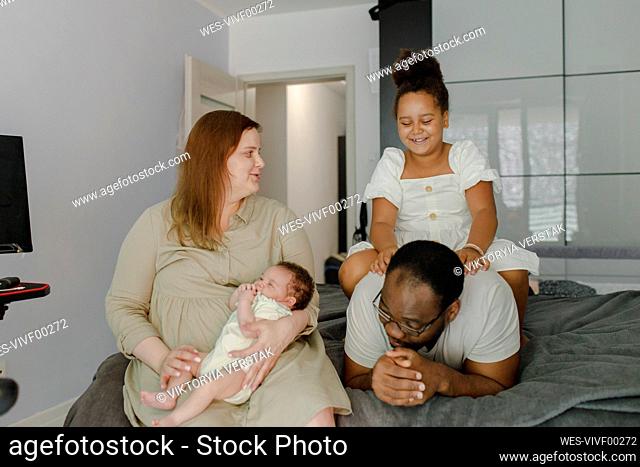 Happy mother looking at daughter sitting on top of her father in bedroom