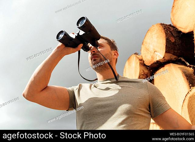 Mid adult man looking through binoculars while standing against sky in forest