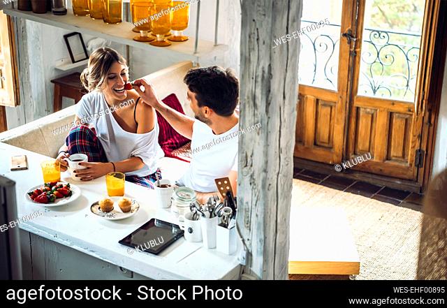 Young man feeding girlfriend with strawberry during breakfast at home