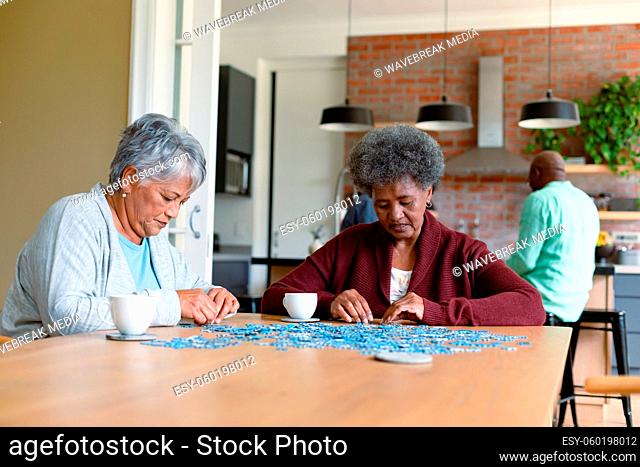 Two diverse female friends sitting in kitchen with coffee and doing puzzles
