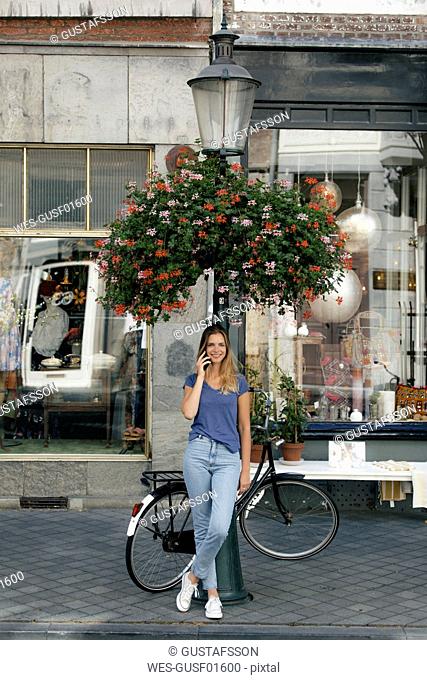 Netherlands, Maastricht, smiling young woman on cell phone at lamp pole in the city