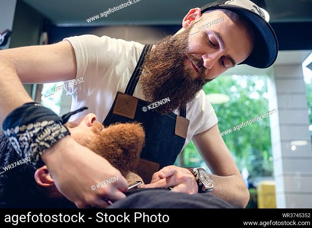 Side view headshot of a redhead bearded young man smiling, ready for shaving in the hair salon of a skilled barber with a classic straight razor in his hand