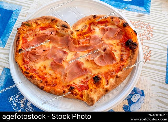 Heart shaped pizza with tomatoes and ham for Valentines Day on white plastic plate on the table, Food concept of romantic love