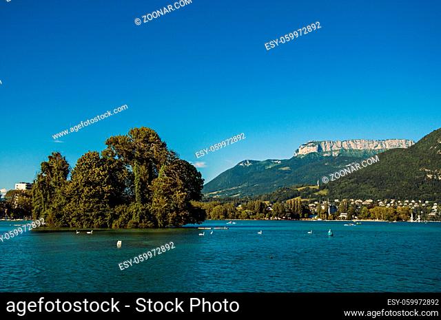 View of Annecy lake, with island, vegetation, mountains and blue sky in background. Located in the department of Haute-Savoie, Auvergne-Rhone-Alpes region