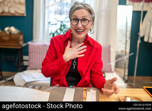 Question. Smiling gray-haired emotional woman in glasses looking questioningly at camera sitting at table with patterns in atelier