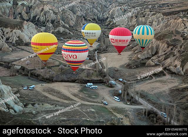 Five multi-colored air balloons are flying above a road in the valley. Balloons colored in red, blue, white, yellow, orange, green