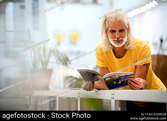 Adult male designer leaning on table while reading magazine
