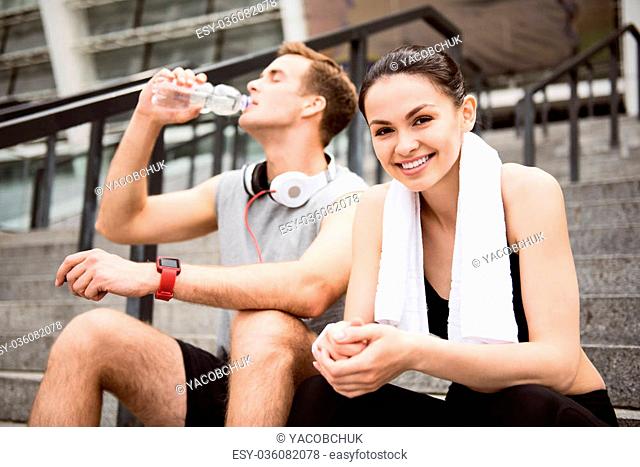 Get some relaxation. Cute young woman looking at camera near a man drinking water while having a rest after exercising