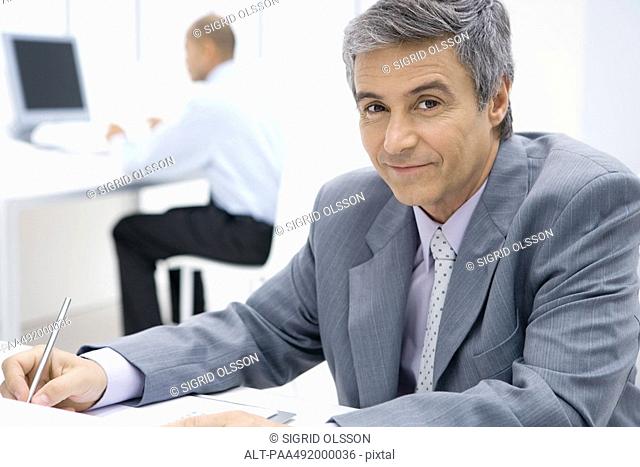 Businessman sitting at desk, using pen, smiling at camera